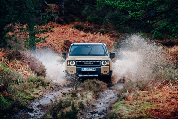 Ein beiger Toyota-SUV fährt durch einen schlammigen Waldweg, spritzt Wasser um sich herum, umgeben von üppigen grünen Bäumen und Herbstlaub.
