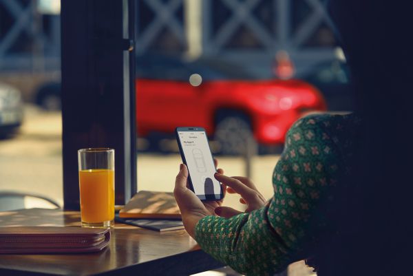 Eine Frau sitzt an einem Restauranttisch und benutzt die MyToyota-App auf ihrem Telefon, ihr Toyota Yaris Cross ist im Hintergrund geparkt.