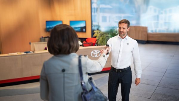 Ein Mann, gekleidet in ein weißes Hemd mit aufgesticktem Toyota-Logo, reicht einer Dame mit ausgestreckter Hand einen Autoschlüssel. Im Hintergrund sieht man einen Empfangsthresen, auf dem das silberne Toyota-Logo hängt.